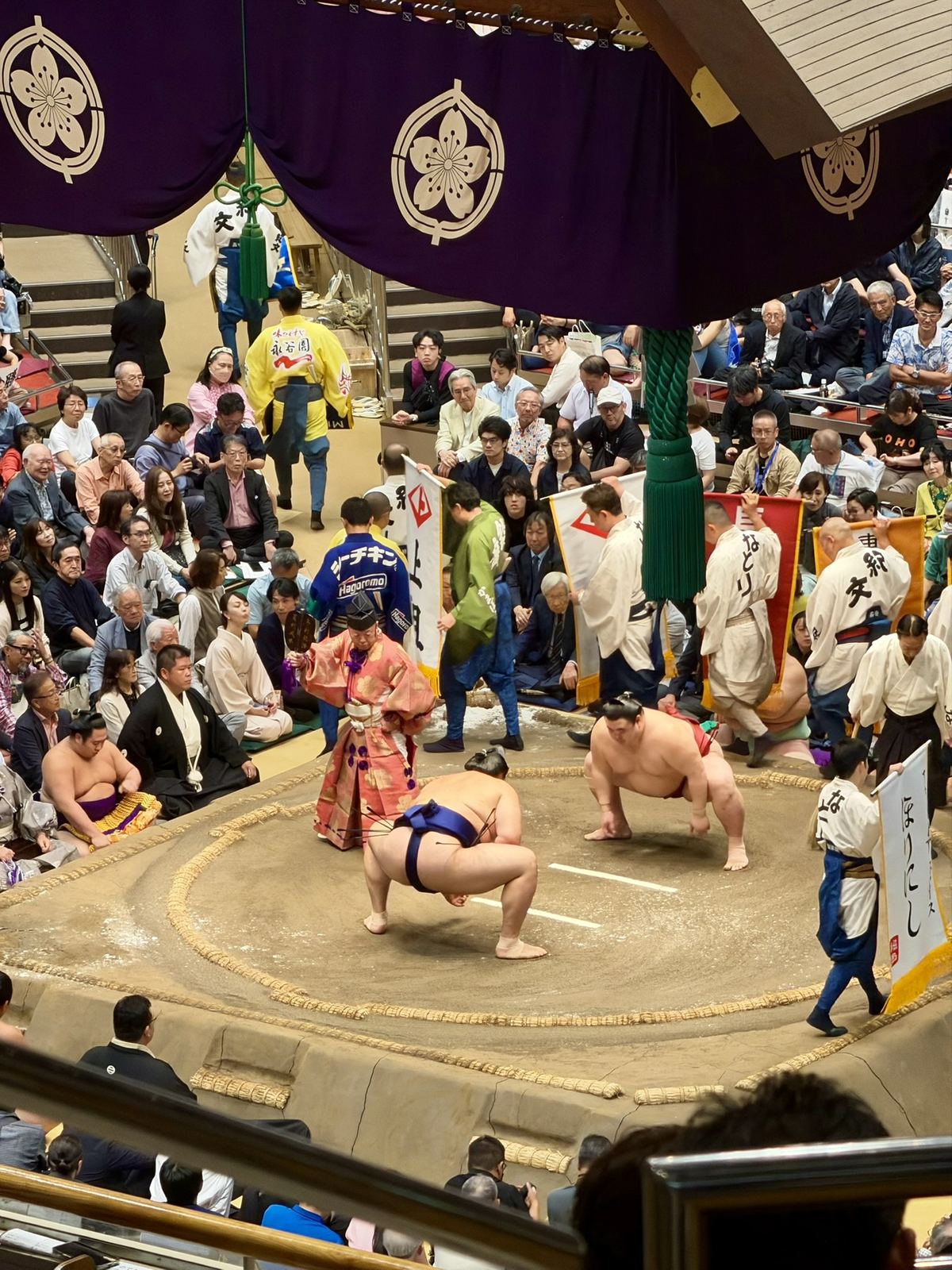取組前に懸賞旗を持って土俵を回る人たち。仕切り中の力士と、オレンジ色の装束の行司も見える