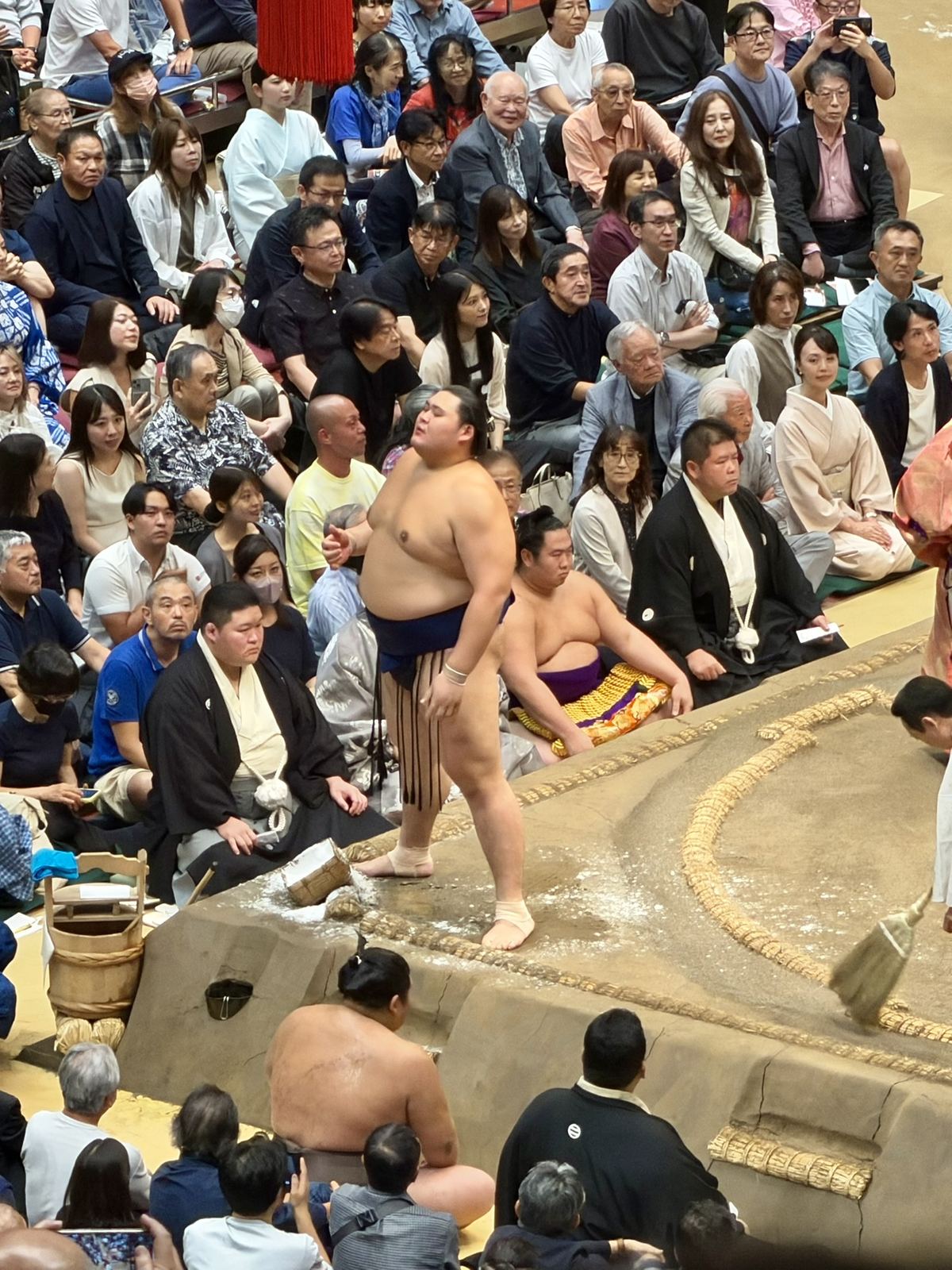 塩かごの横、土俵際に立つ力士。後ろには観客と黒紋付の審判がいる