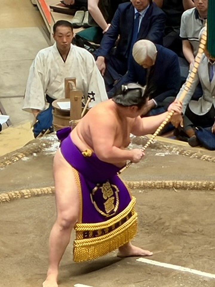 紫の化粧廻しを締めた力士が、土俵の上で長い弓を振っている
